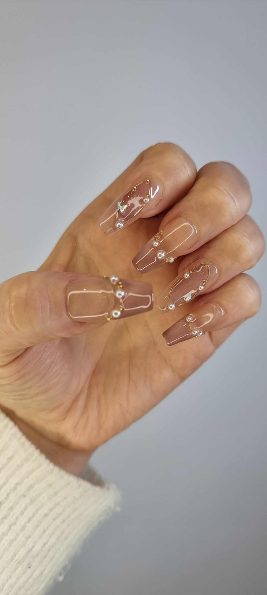 Hand wearing Khawla long coffin press-on nails in brown and nude, with detailed gold chain and pearl embellishments.