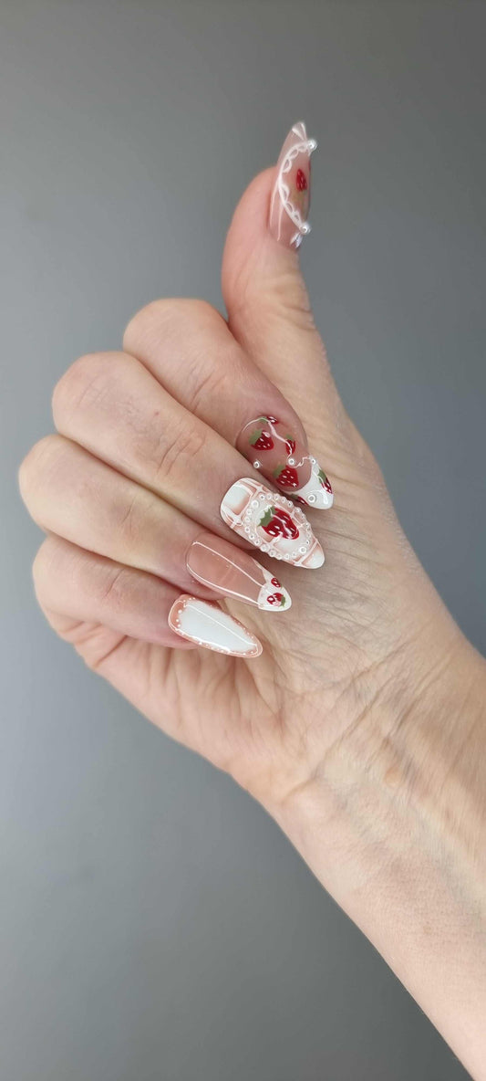 Omani Oasis press-on nails in a long almond shape, featuring a charming hand-painted strawberry design with white lace and pearl details on a nude base.