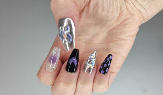 A set of silver, glossy, and black press-on nails. Some nails feature a holographic liquid-like design, while others have black abstract art, purple glitter, or a black and white checkerboard pattern.