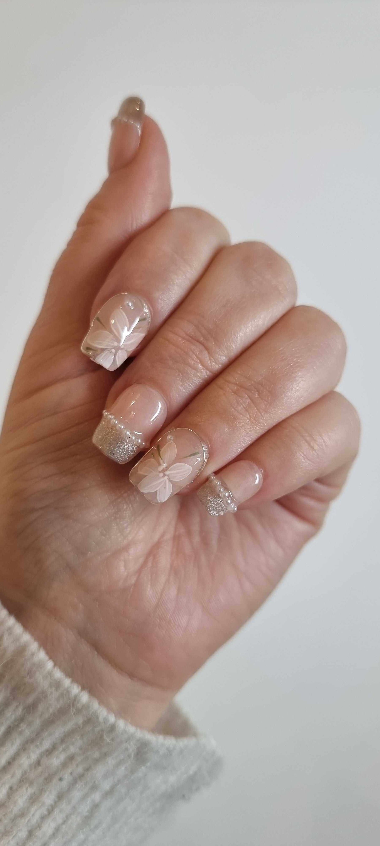 Hand with elegant beige nail polish featuring white floral designs and glittery tips.