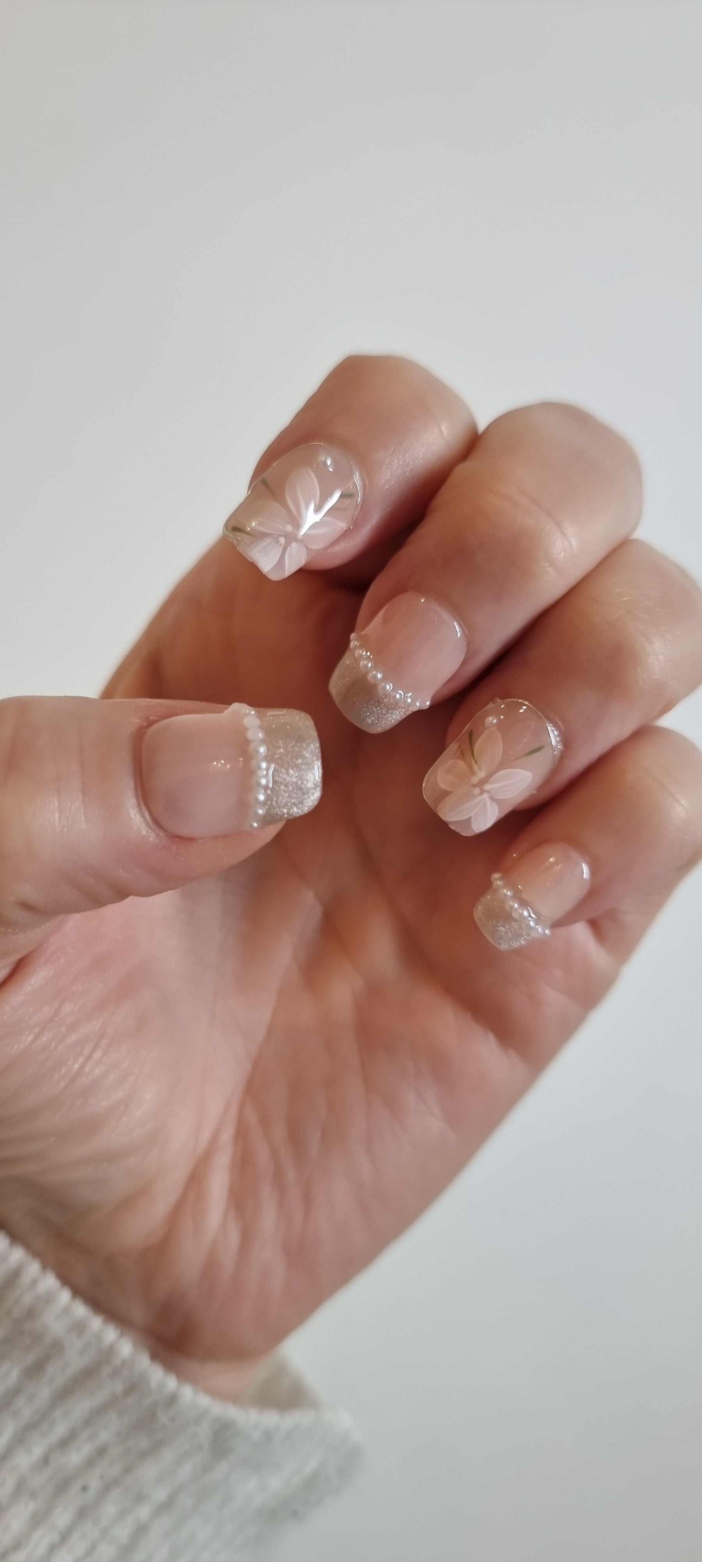 Close-up of manicured hand with elegant, natural-toned nails featuring delicate white floral and glitter designs, showcasing Zohra nail gel style.