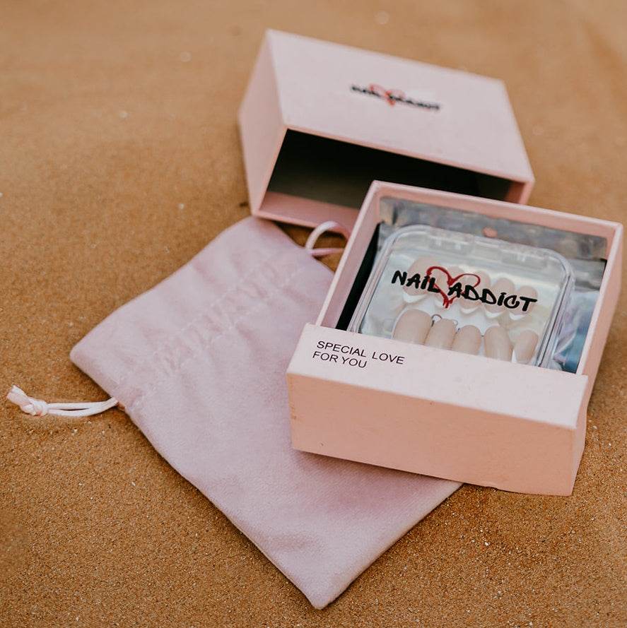 A luxury Nail Addict press-on nail set in its pink box and velvet pouch, displayed on sand, representing the best place to buy reusable press-on nails in Abu Dhabi.