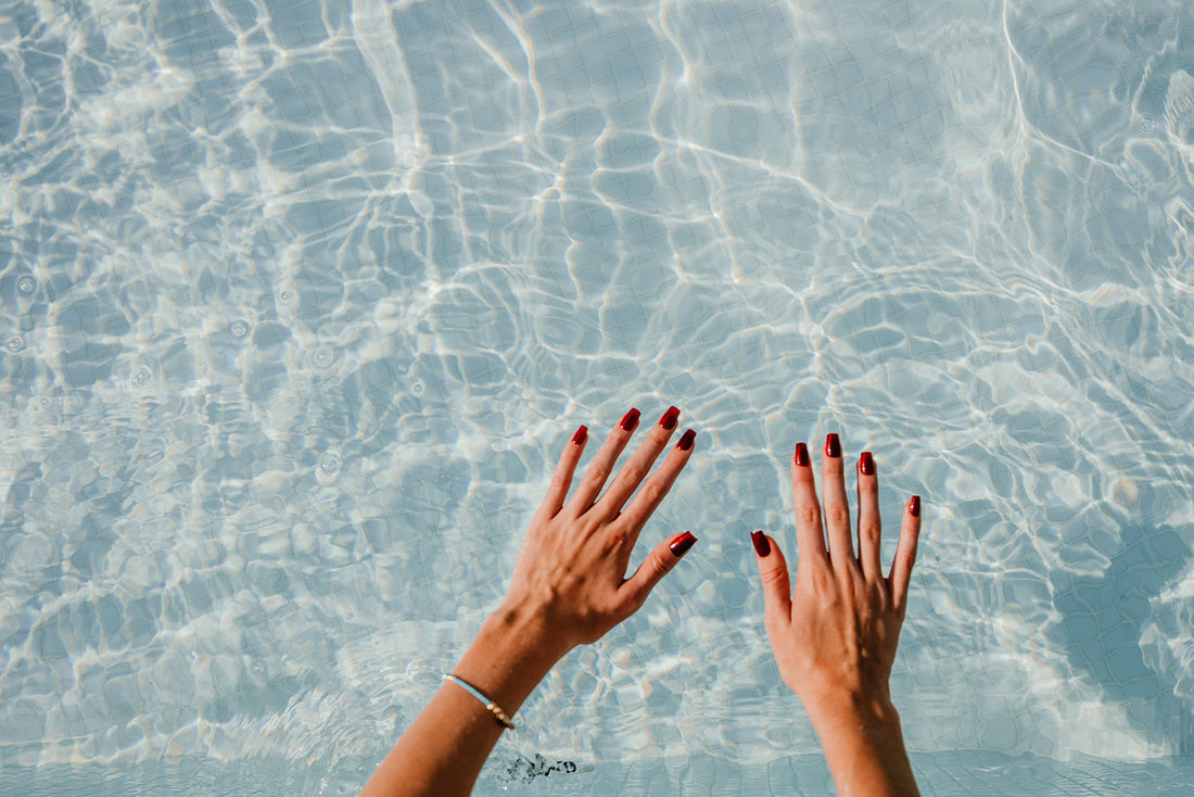 A woman's hands with a fresh red manicure floating in clear blue water, representing the top spring nail trends for 2025.