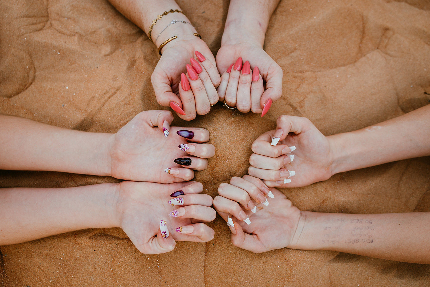 Group of hands displaying a variety of manicures including classic coral pink and decorative white nail art against a natural sand background, showcasing professional nail products in the UAE.