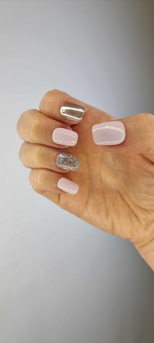 A hand with a full set of five short, square-shaped press-on nails. The thumb is a solid silver chrome, the ring finger has silver glitter, and the other three fingers are pale pink.