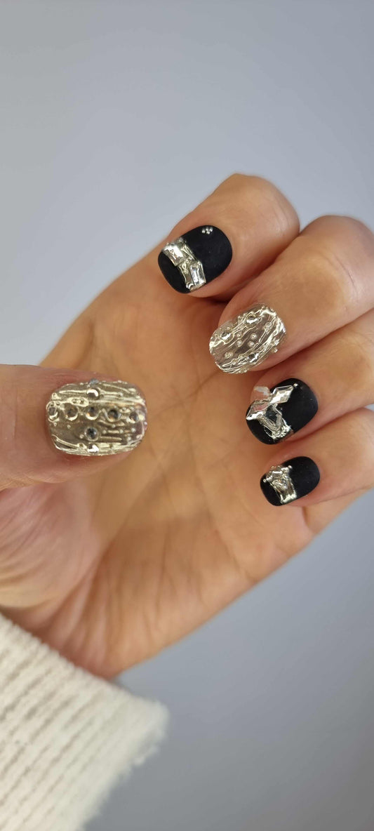 A set of black and silver glossy press-on nails. The design includes a mix of black solid colors, a silver abstract swirl pattern, and intricate rhinestone details on a textured background.
