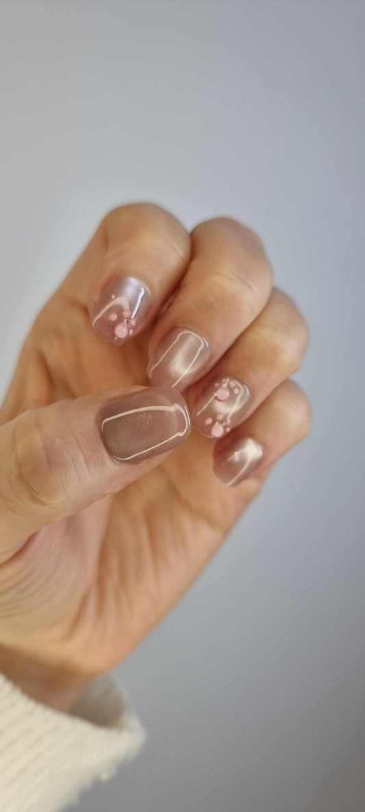 A set of glossy, neutral-toned press-on nails. Some nails feature a subtle pearlescent sheen, while others have a small paw print pattern in a darker nude shade.