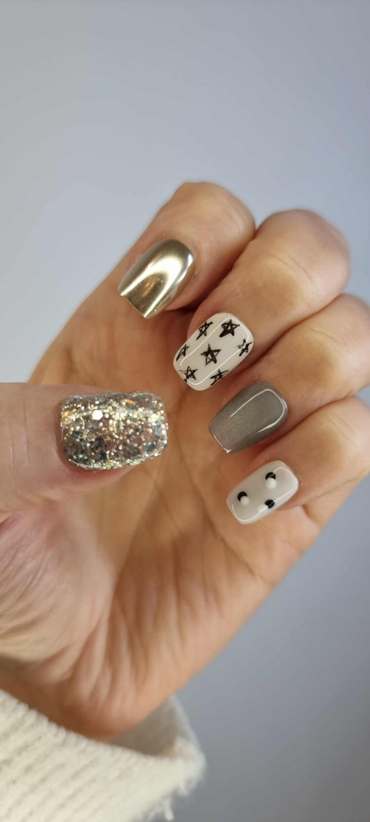 A set of glossy, neutral-toned press-on nails. Some nails are covered in silver glitter, while others feature a metallic gold and silver celestial design with moons and stars.