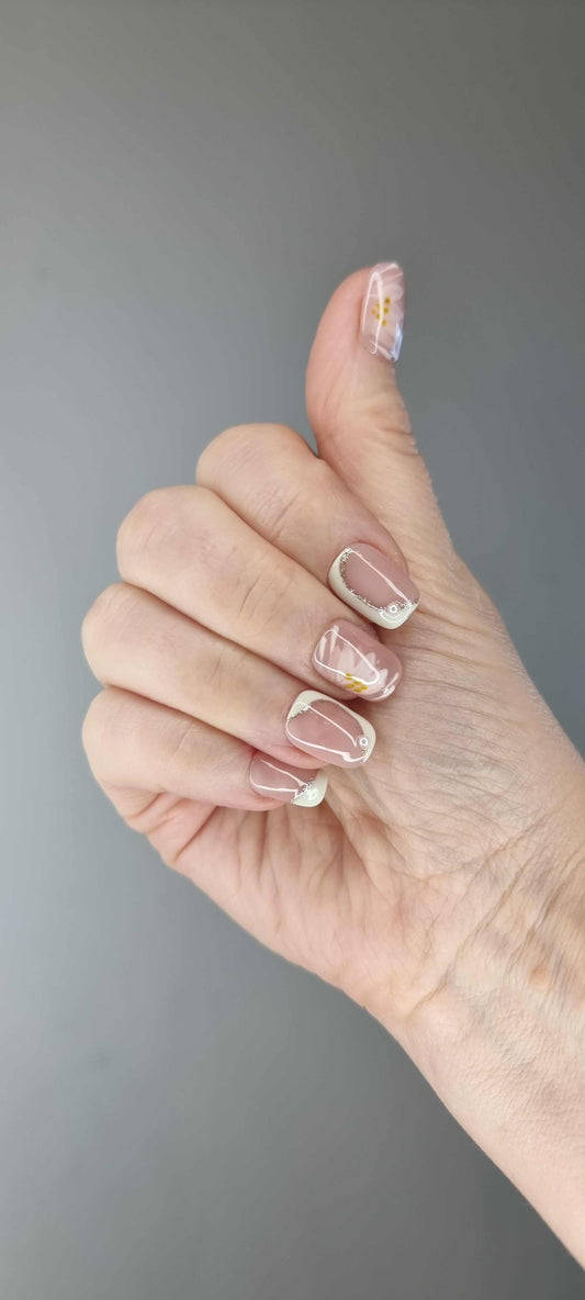 A set of false nails with a nude beige base. The nails feature a soft white and gold floral design, resembling daisies, with small gold accents.