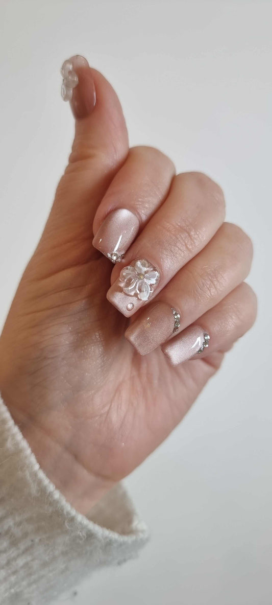 A set of false press-on nails in a glossy, light brown or nude tone. The nails are decorated with delicate white 3D floral shapes and tiny rhinestone details.