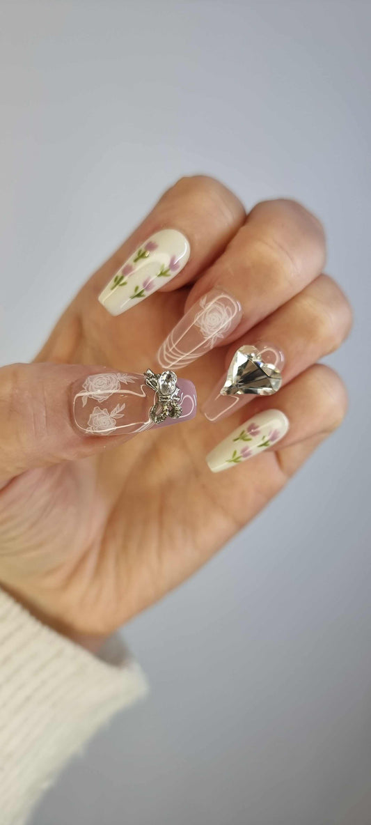 A set of light, neutral-toned press-on nails. Some nails feature small red and yellow floral designs, while others are decorated with large clear rhinestones and silver details.