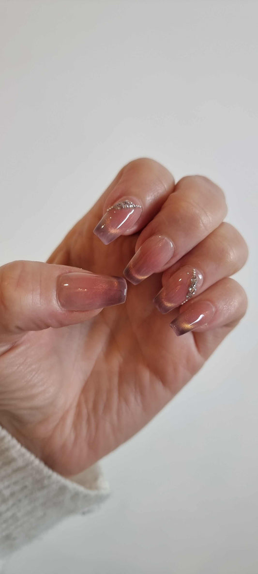 A set of long, square-shaped press-on nails with a glossy brown and beige swirled design, featuring a diagonal line of rhinestones on some of the nails.
