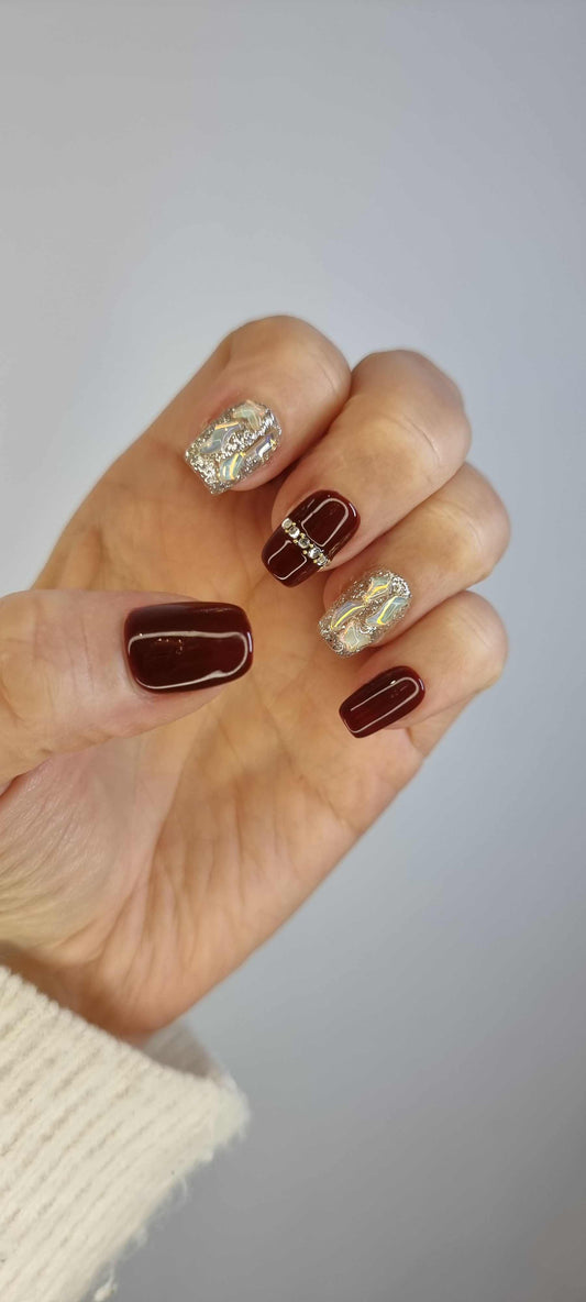 A set of red, short square press-on nails. Some nails are a solid glossy red, while others are covered in a gold glitter design with rhinestone bands in the middle.