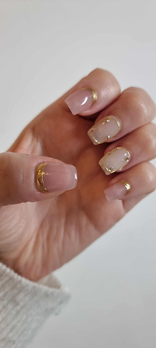 A set of nude artificial nails with a glossy finish. The nails feature a metallic gold-painted French tip and small gold rhinestone details.