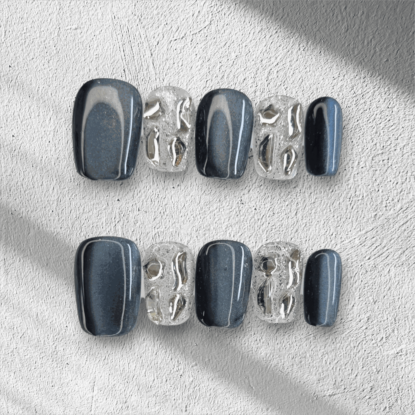 The Days in Dubai press-on nail set in a short square shape, featuring a mix of grey-blue and textured clear nails with molten chrome details.