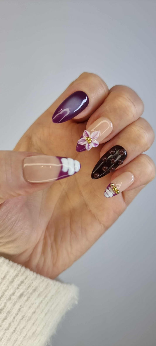 A model's hand wearing the elegant Emirati Essence press-on nails, showcasing the intricate purple and gold floral designs.