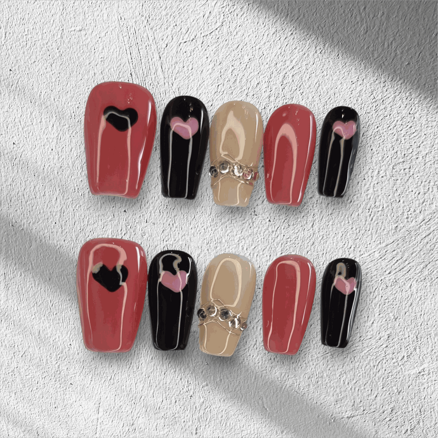 The Hind press-on nail set in a long coffin shape, featuring a mix of red, black, and nude nails with heart designs and rhinestone chain accents