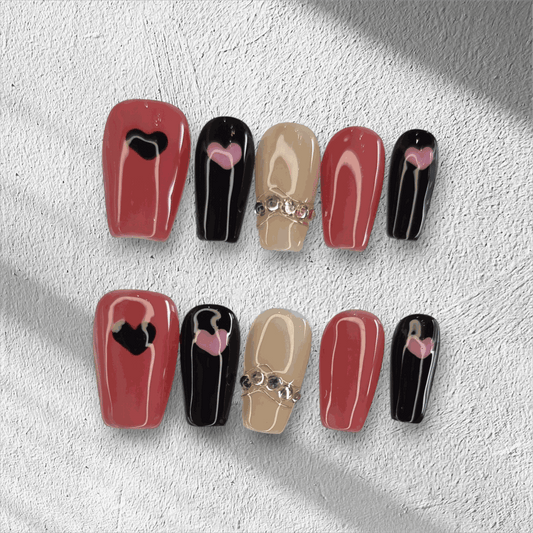The Hind press-on nail set in a long coffin shape, featuring a mix of red, black, and nude nails with heart designs and rhinestone chain accents