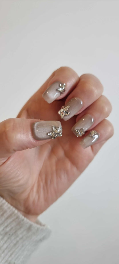 Hand wearing Jeddah Glow pearlescent nude press-on nails, showcasing the unique 3D silver flower and leaf charm on the ring finger.