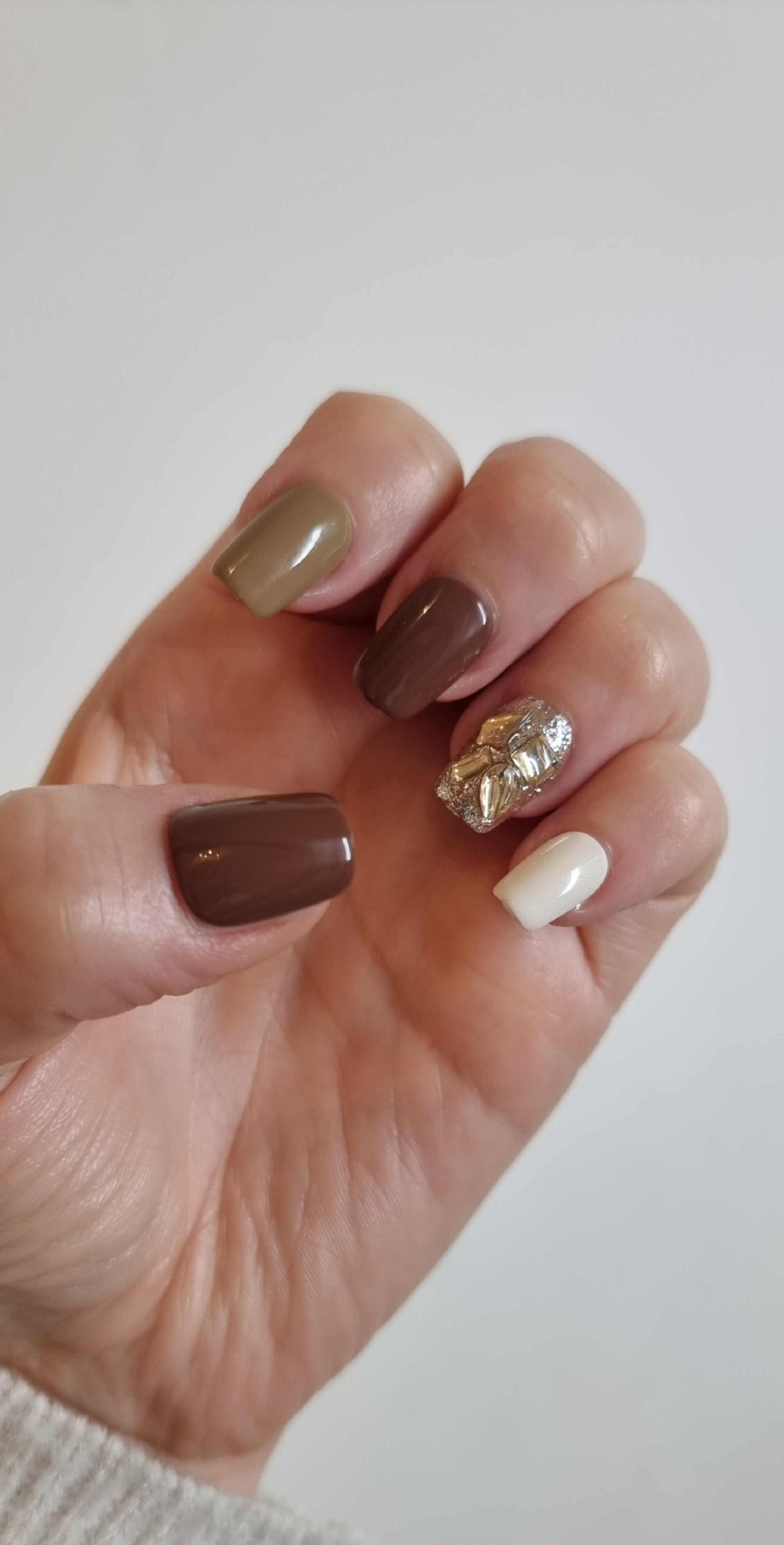  Hand wearing Jumeirah Glow short square press-on nails in various earth tones with a gold foil and rhinestone design on the ring finger.