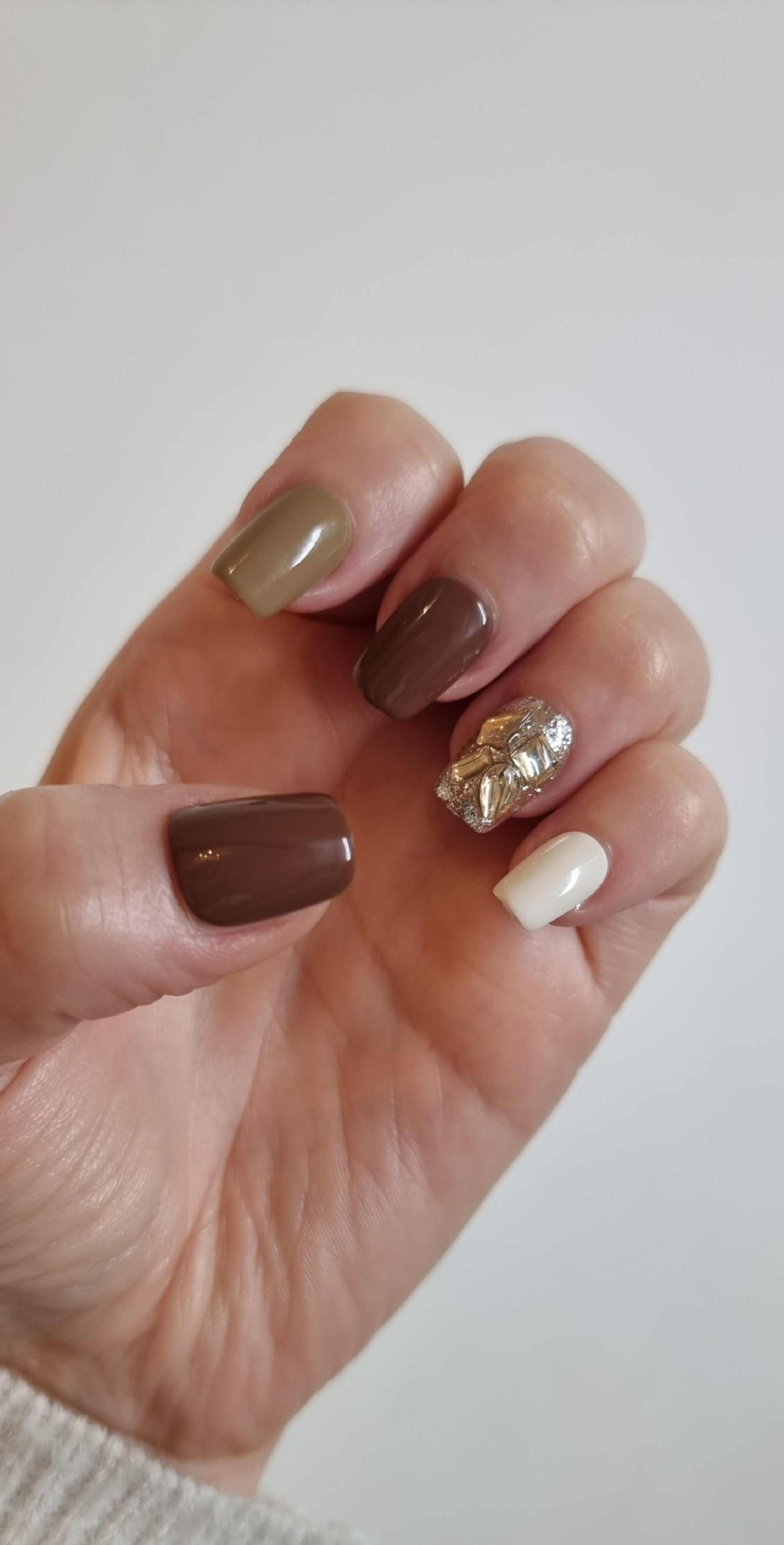  Hand wearing Jumeirah Glow short square press-on nails in various earth tones with a gold foil and rhinestone design on the ring finger.
