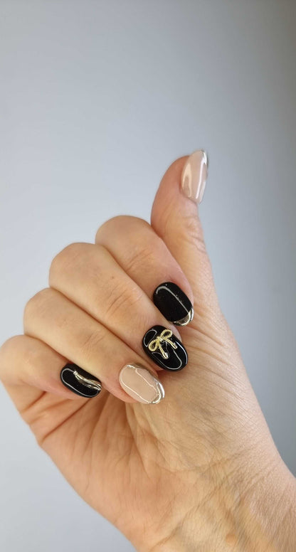 Hand wearing Muscat Mystique black and nude almond press-on nails with elegant gold bow and metallic accents.