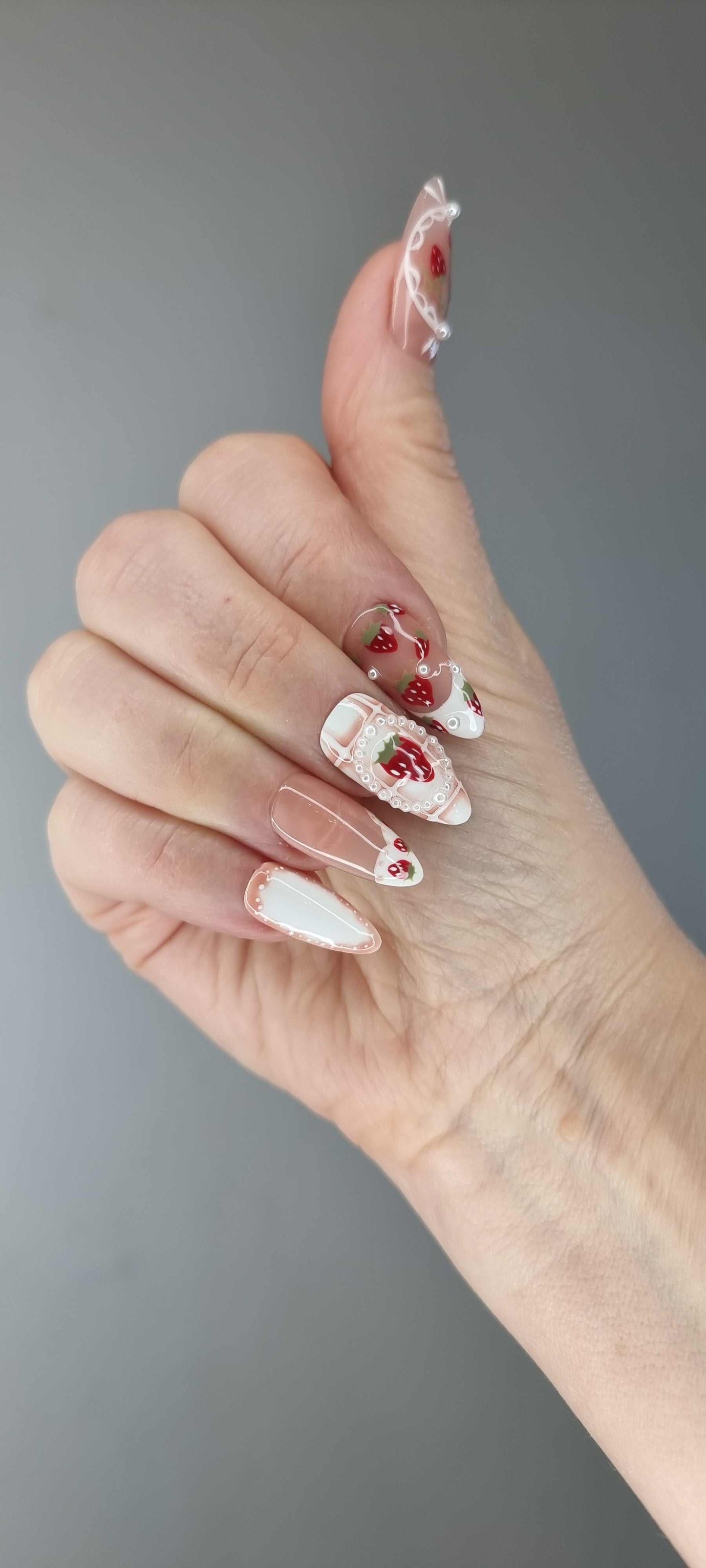 Omani Oasis press-on nails in a long almond shape, featuring a charming hand-painted strawberry design with white lace and pearl details on a nude base.