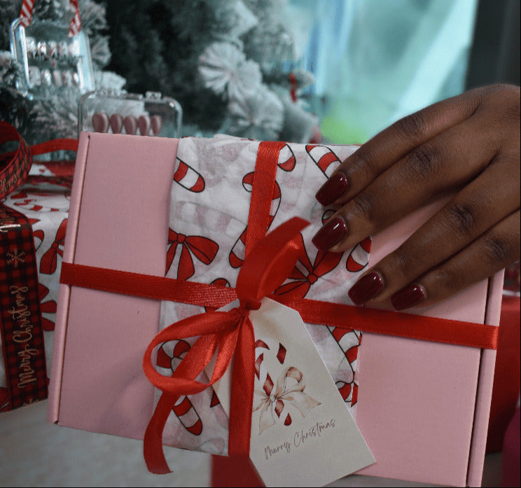 Hand holding a pink gift box with a red ribbon and candy cane design, Christmas tree in the background
